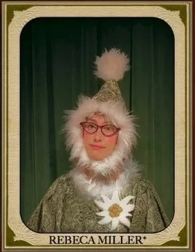 Woman wearing festive winter hat with pom-pom and fur, glasses, holding a white flower, standing against a green curtain background.