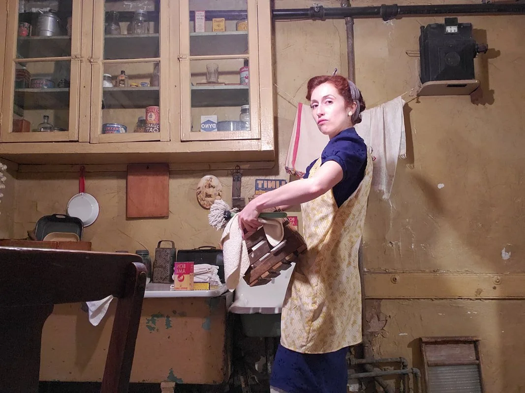 A woman wearing a vintage-inspired dress and apron standing in a cluttered kitchen, holding a tray and a dish towel.
