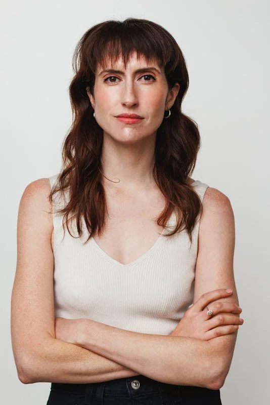 A woman with shoulder-length brown hair, wearing a sleeveless white top, standing with arms crossed against a plain white background.