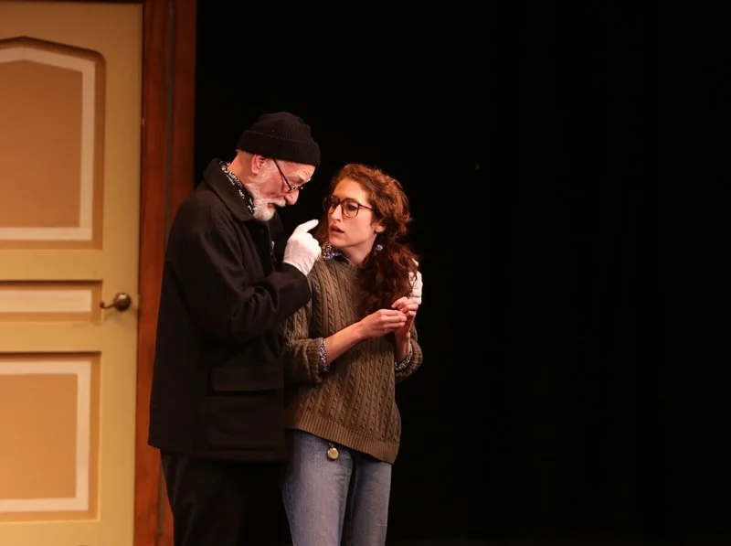 Two actors performing on stage; an older man with glasses, black beanie, black coat, and white gloves is speaking to a younger woman with glasses, long curly hair, a brown sweater, and jeans, next to a door.