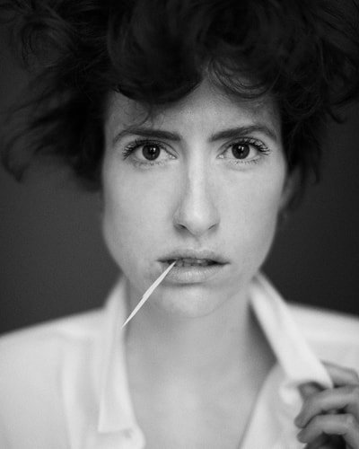 Black and white portrait of a woman with short, curly hair holding a toothpick in her mouth and looking directly at the camera.