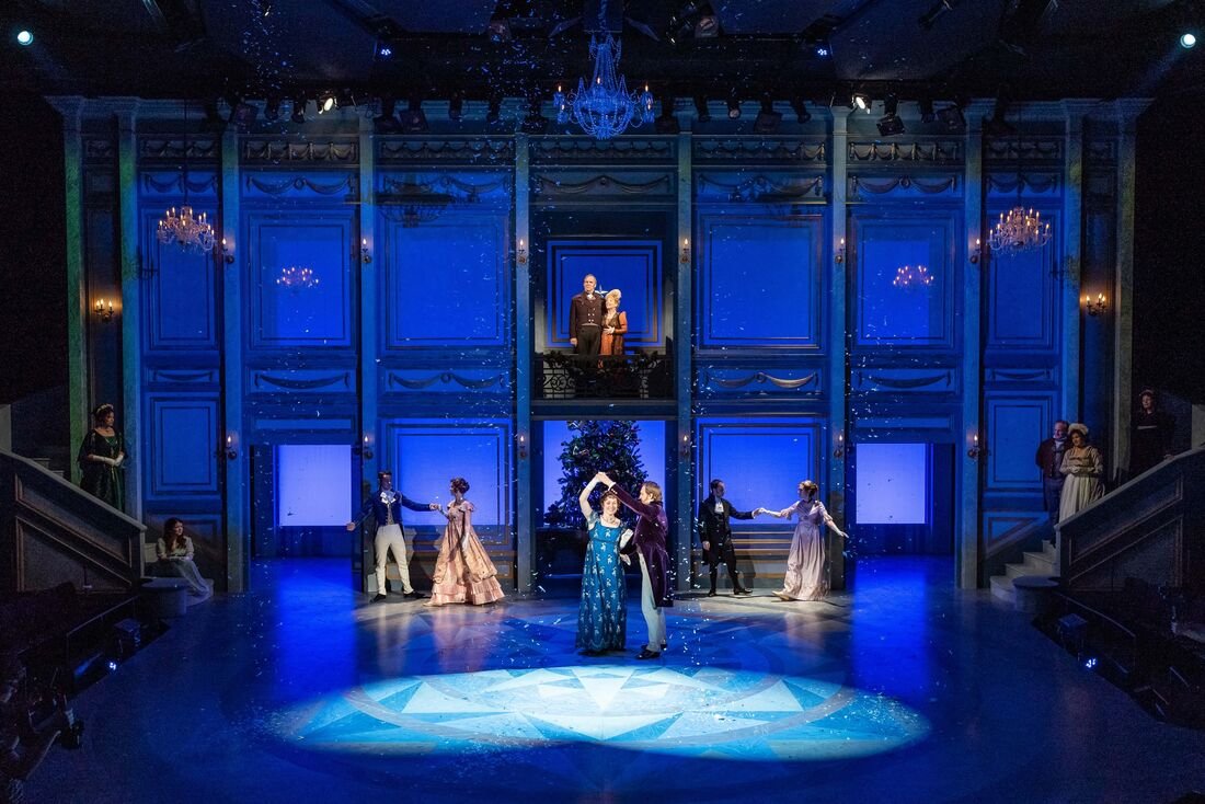 A theatrical stage with elaborate blue decor, chandeliers, and multiple performers in period costumes. Two dancers are center stage, with one in a blue gown and the other in a dark suit, dancing under a spotlight. Other performers are on the stage an