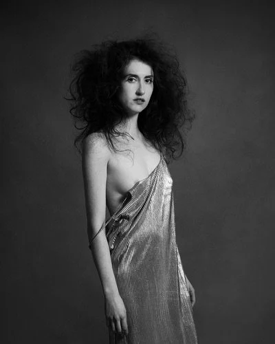 A woman with curly hair wearing a shiny, sleeveless dress, standing against a dark background.