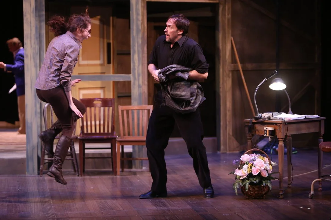 A woman with curly hair and glasses, wearing a gray jacket and brown boots, appears to be jumping or in an animated pose, while a man with dark hair and a beard, dressed in black, holds a coat and faces her. They are on a wooden stage with chairs, a 