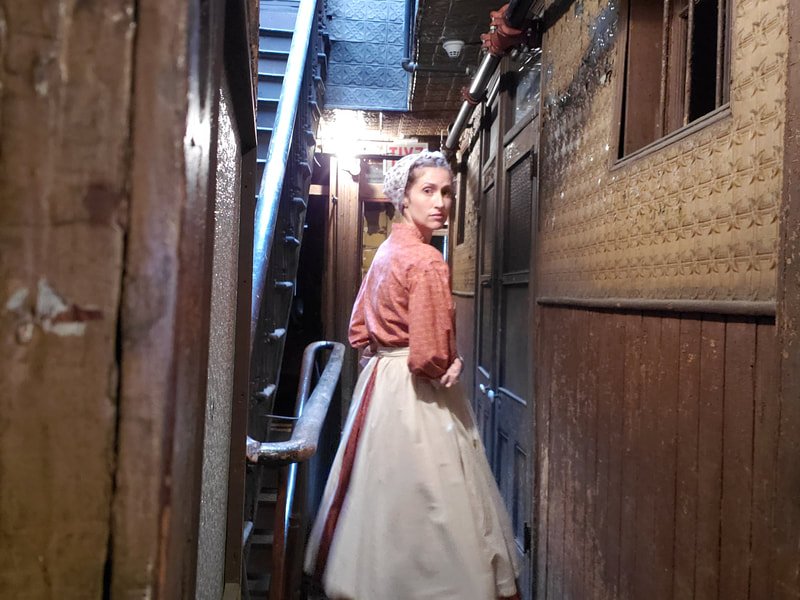 A woman wearing a 19th-century dress with a cream-colored skirt, a reddish-brown blouse, and a white bonnet, standing in a narrow, dimly-lit, rustic hallway with old wooden walls and a metal staircase.