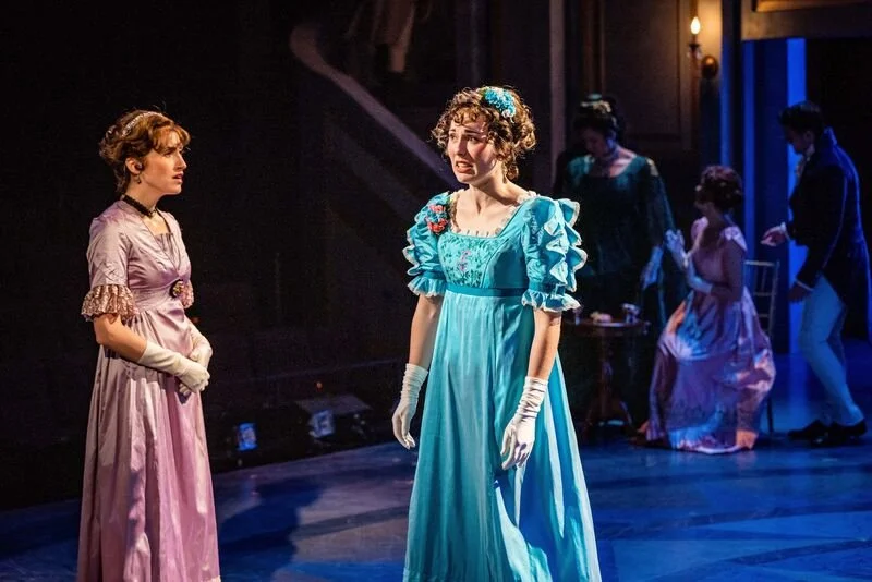 Stage scene from a play or musical set in historical period, featuring women in elaborate 19th-century dresses with ruffled sleeves and gloves, engaged in discussion.