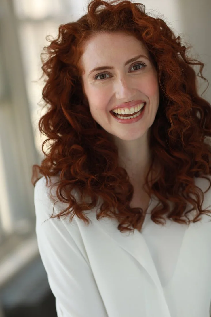 A smiling woman with long, curly red hair wearing a white blouse, standing indoors near a window.