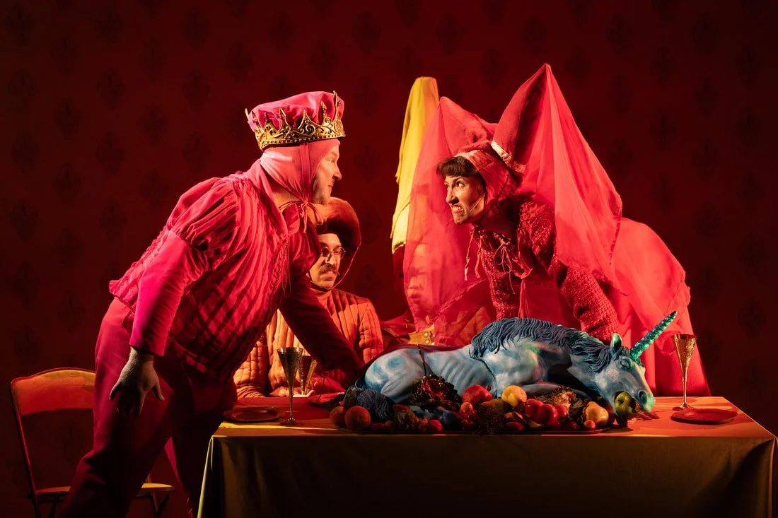 The image shows a theatrical scene with three men in elaborate costumes around a table with a cake shaped like a unicorn. One man, wearing a crown and red robe, leans forward aggressively. The second man is dressed in red, including a pointed hat, an