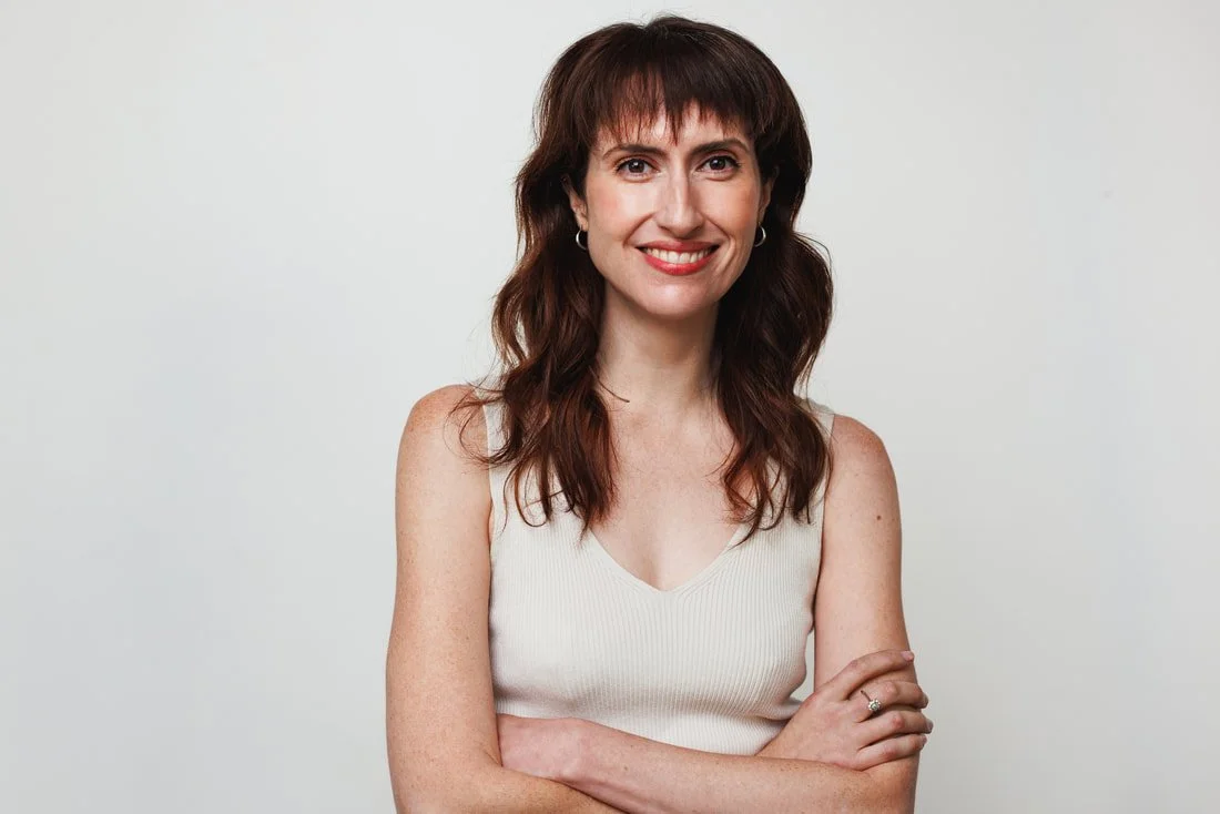A woman with brown, wavy hair, wearing a sleeveless cream top, smiling with arms crossed against a plain light gray background.