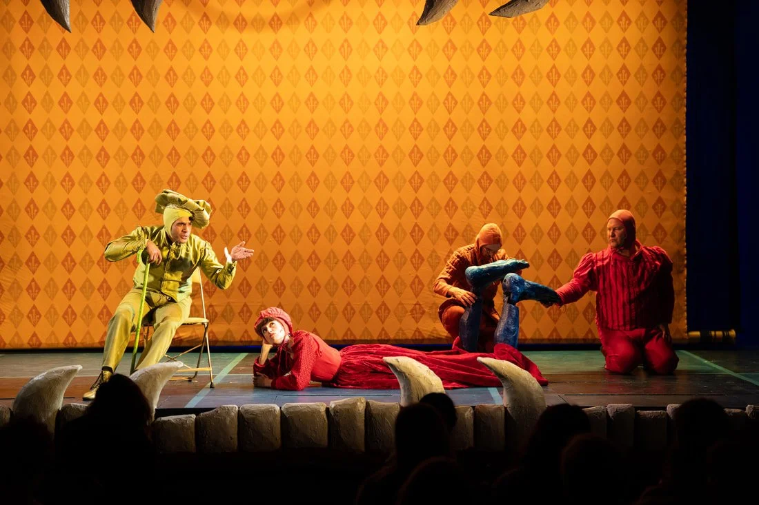 The stage features actors in colorful costumes performing. One actor in a yellow costume is sitting on a chair gesturing with animated expressions. A woman in red with a pink hat is lying on the floor, looking up. Two actors, one in orange and anothe