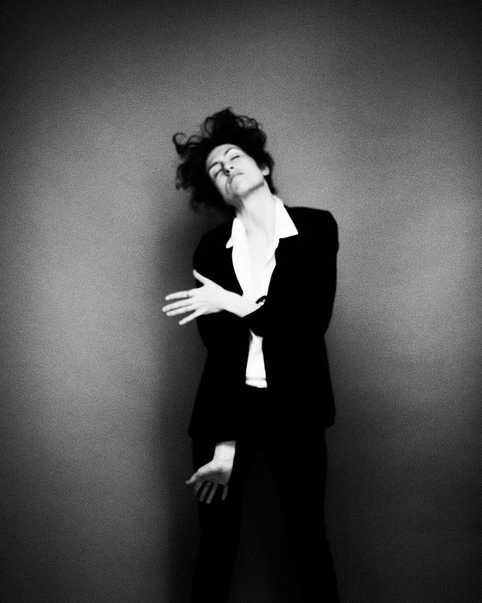 Black and white photo of a woman with curly hair, wearing a suit and white shirt, standing against a plain background. She appears to be in a relaxed, artistic pose with her eyes closed and head tilted back.