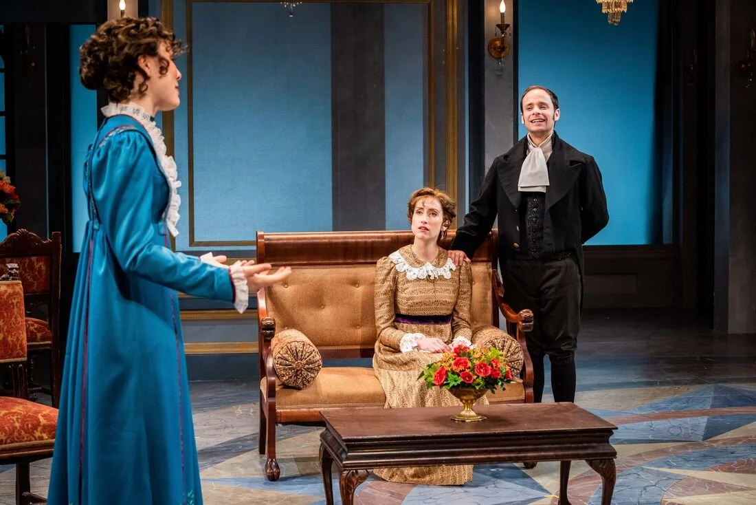 A theater scene with three actors. A woman in a blue period dress stands on the left, speaking to a woman seated on a vintage sofa, who has a bouquet of red flowers on her lap. A man stands on the right, smiling and resting his hand on the woman's sh