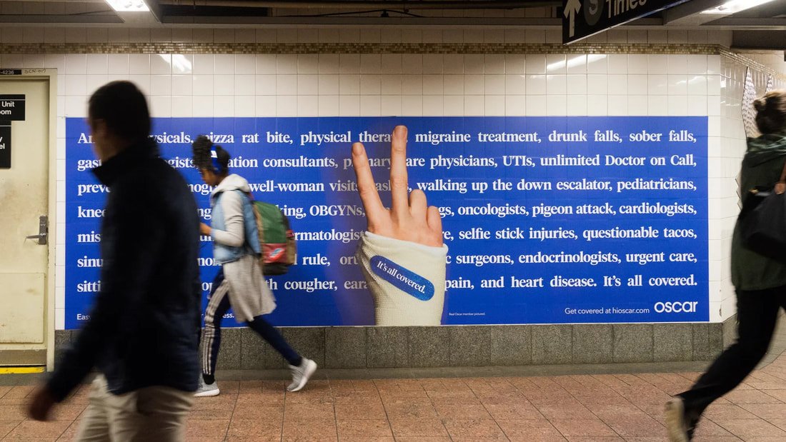 Large blue advertisement billboard in a subway station with a hand giving a peace sign. The billboard contains text about health services and has a sticker that reads 'It's all covered.' Several people are walking past the billboard.