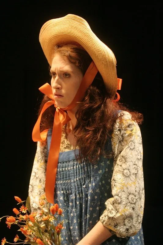 A woman dressed in vintage clothing with a large straw hat, orange ribbons, and a floral dress, holding a bouquet of orange flowers, standing against a dark background.