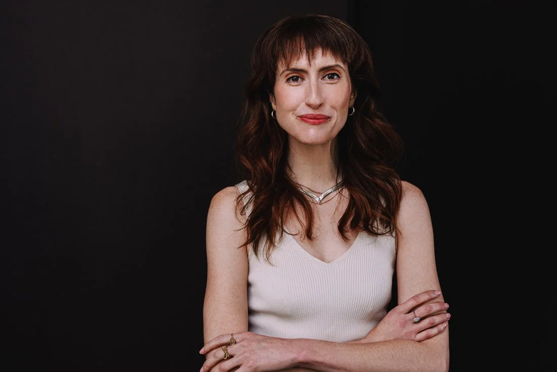 Portrait of a woman with shoulder-length dark brown hair, wearing a white sleeveless top, against a black background.
