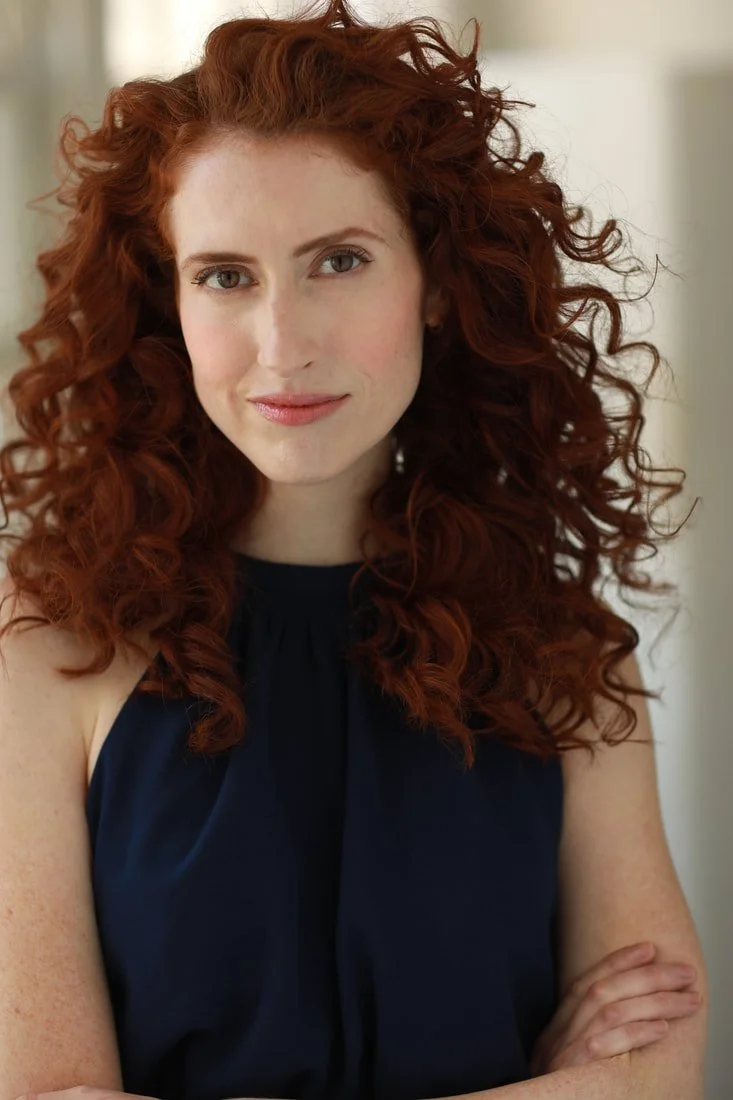 A woman with long, curly red hair wearing a dark sleeveless top, standing indoors with arms crossed, smiling softly.