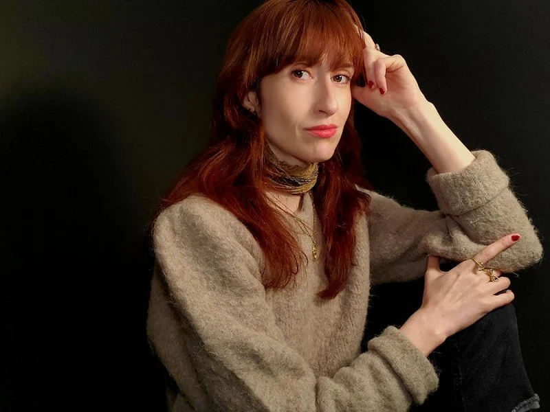 A woman with red hair wearing a beige sweater, a black and gold neck scarf, and gold jewelry, posing against a black background.
