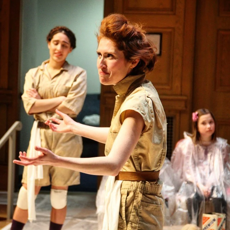 Three women and one young girl in a room with wooden paneling in the background. The woman in the foreground has short, curly red hair and is wearing a tan jumpsuit, gesturing with her hands. The woman in the background has dark hair tied back, weari