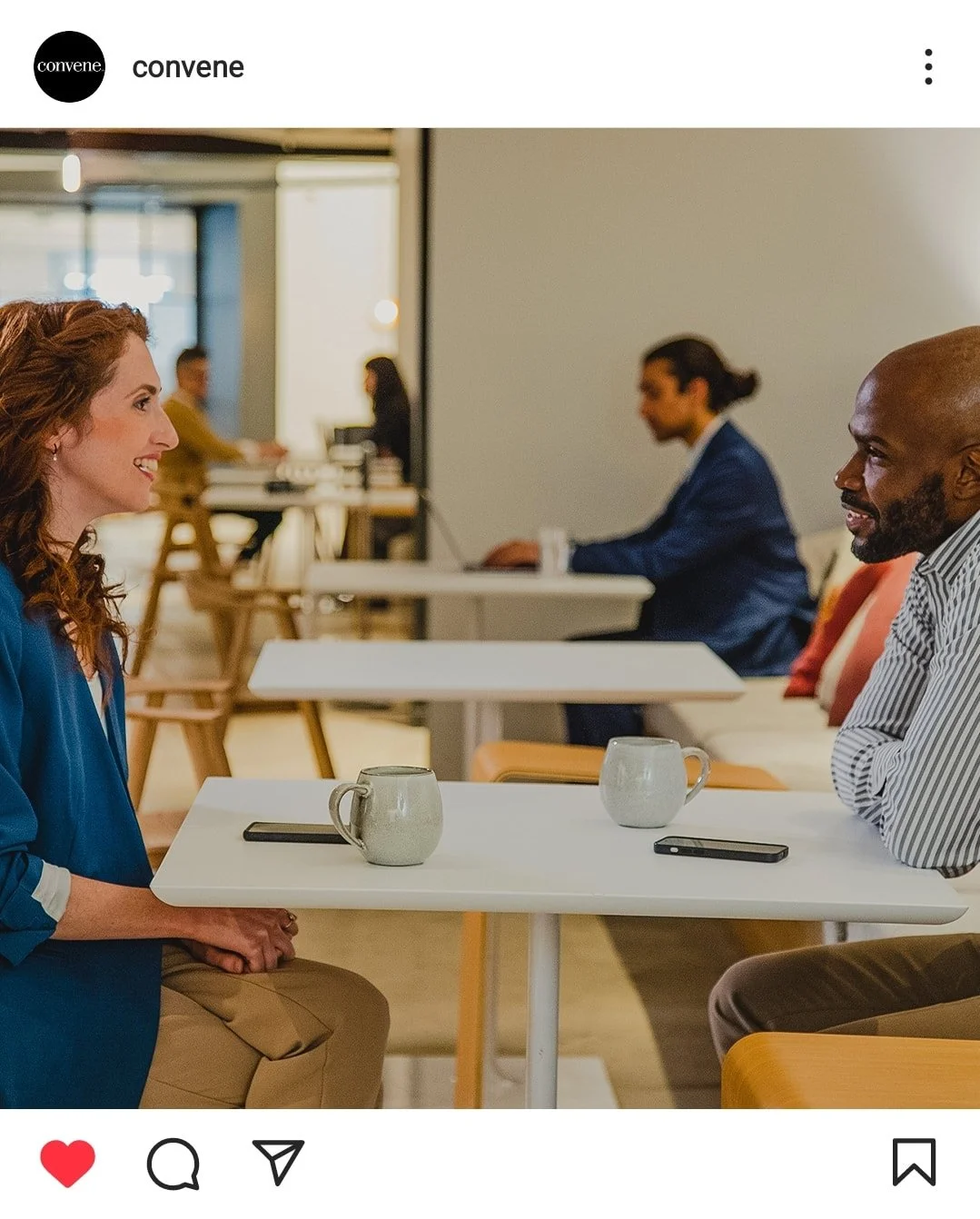 Two people sitting across from each other at a cafe table, smiling and engaging in conversation, with coffee mugs and smartphones on the table.