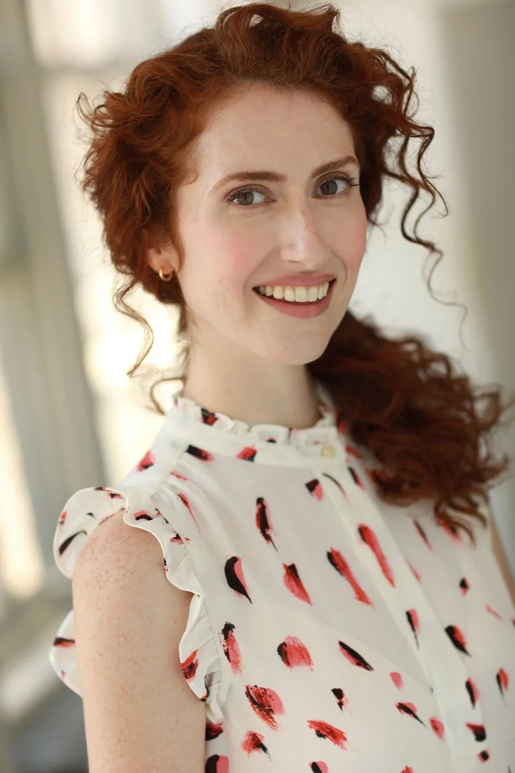 A woman with curly red hair and light skin, smiling, wearing a white blouse with ruffled sleeves and a red and black abstract pattern, standing in front of a neutral background.