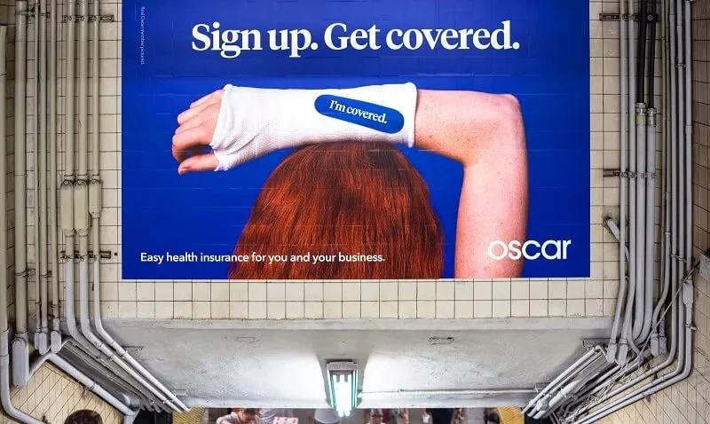 A billboard advertisement for Oscar health insurance featuring a woman with red hair and a white arm cast, promoting signing up for coverage.