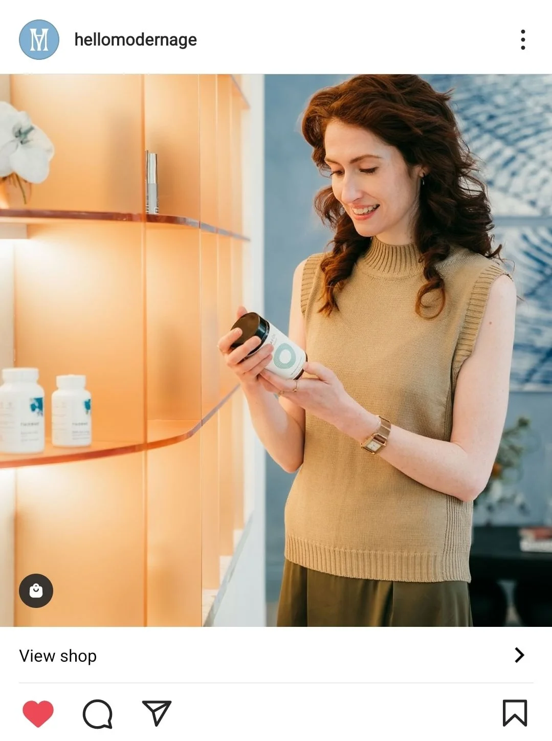 A woman with curly brown hair wearing a beige sleeveless sweater looks at a bottle of supplements or medication in her hands.