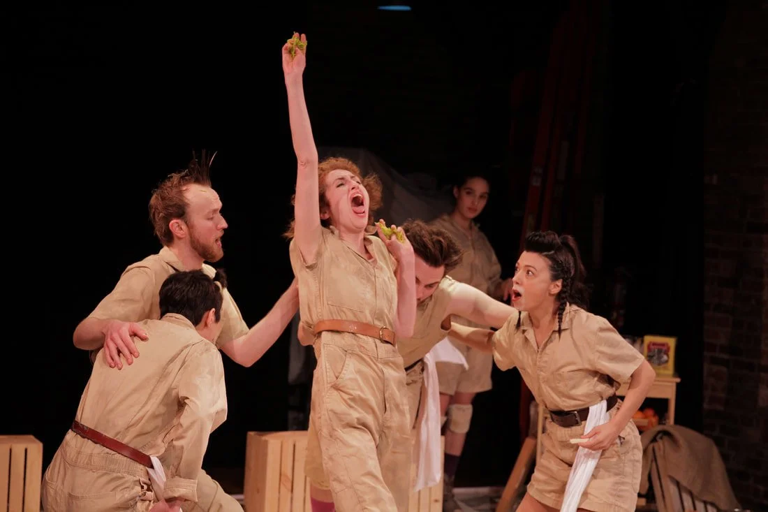 Actors performing a scene on stage, dressed in matching beige costumes, with one actor raising their arm and others reacting dramatically.