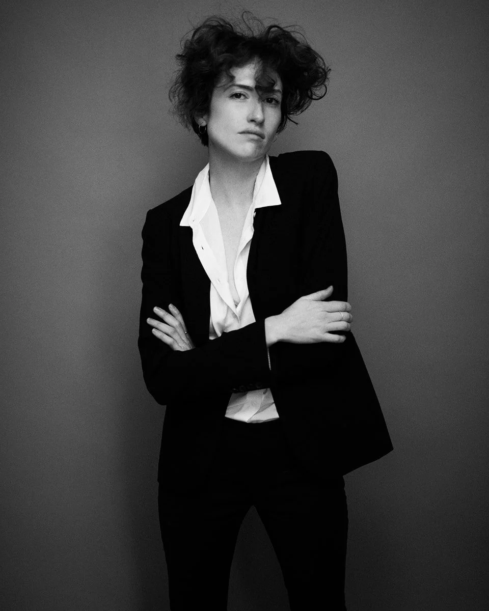 A woman with curly hair wearing a black blazer and white shirt, posing with crossed arms against a plain background in black and white.