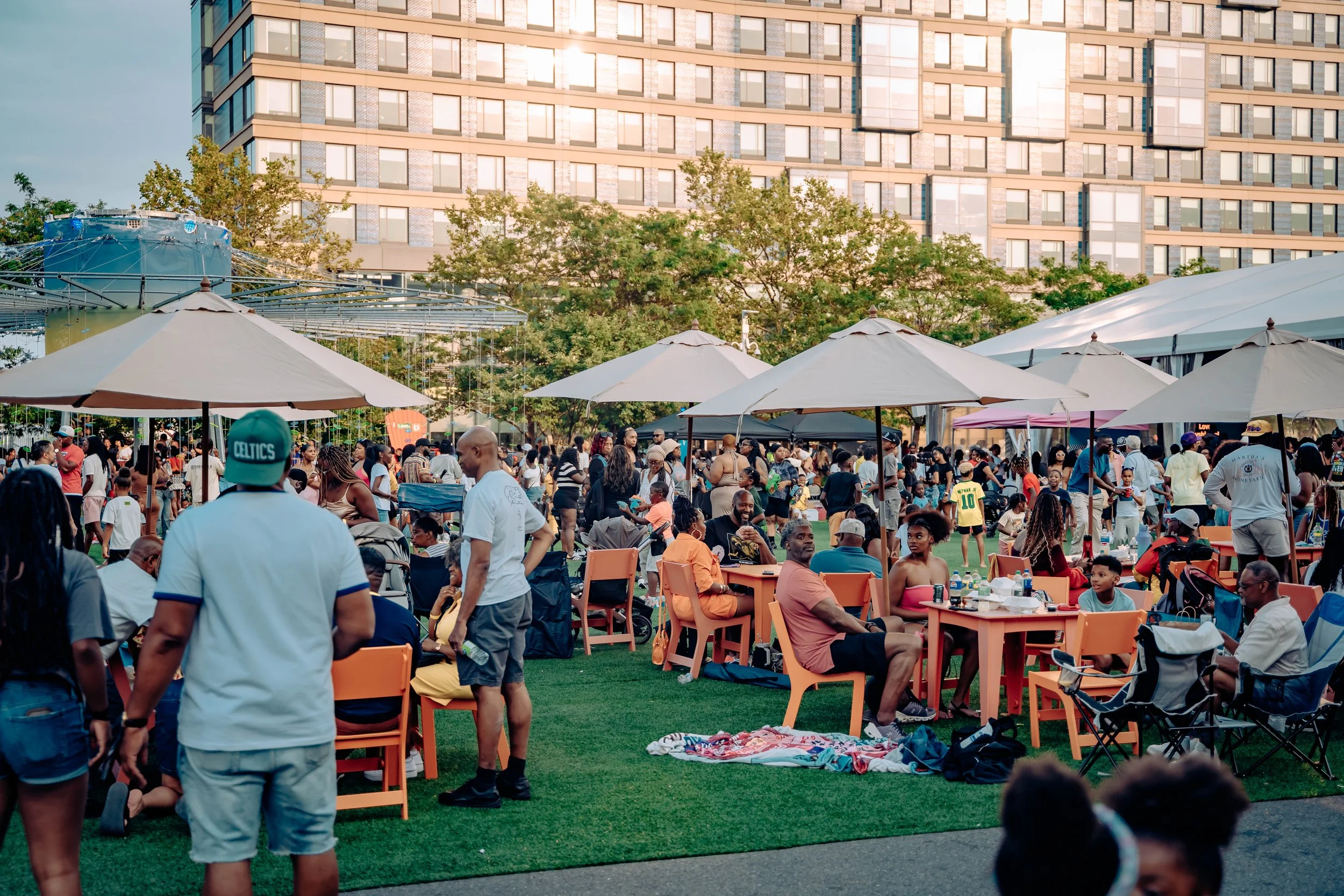 Outdoor gathering of diverse people under umbrellas, sitting and walking on green grass, with a city building in the background.
