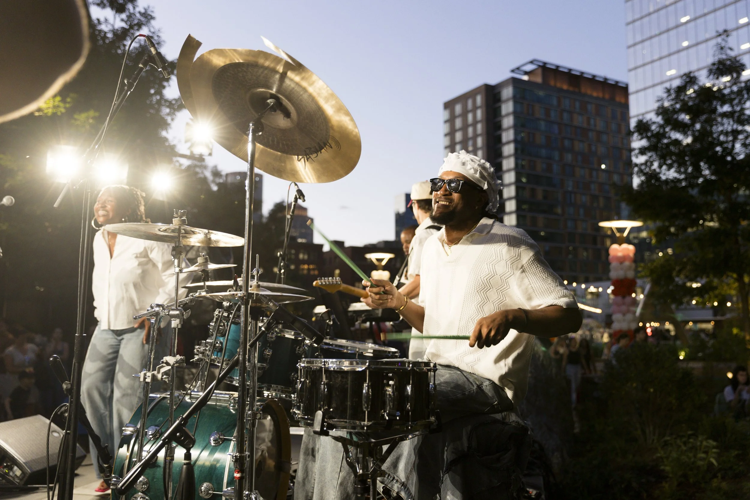A jazz band performing outdoors at dusk in a city park. The drummer is smiling and wearing sunglasses, with a cymbal and drum set in front. Behind him, a woman singer and other musicians are visible. Tall buildings and trees are in the background, and stage lights are shining.