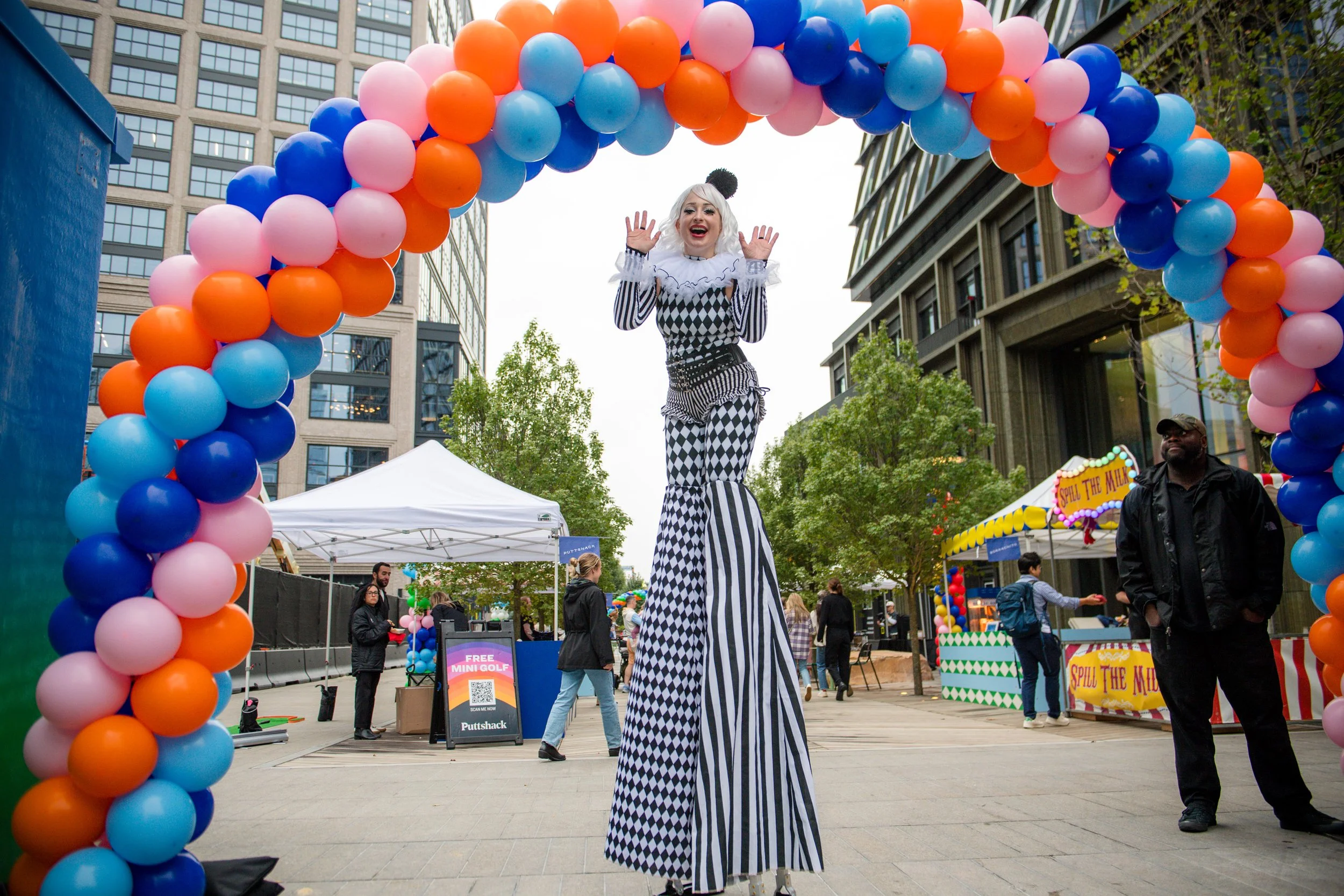 A performer on stilts dressed as a clown with black and white checkered and striped costume, white wig, and black pom pom, stands under a colorful balloon arch at an outdoor fair in a city. People walk past, tents and booths are visible in the background.