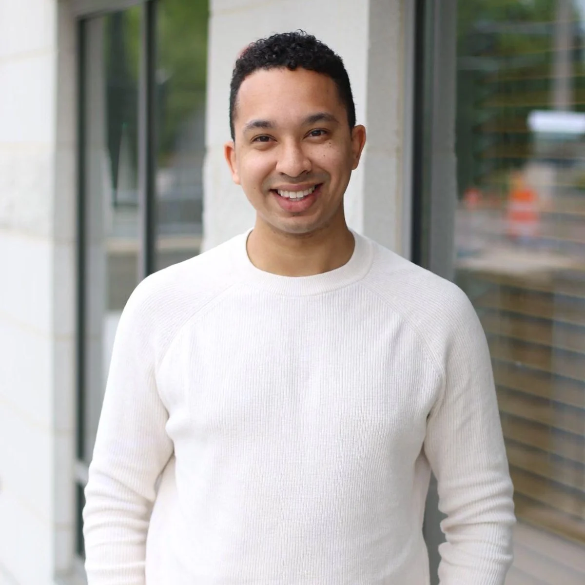 A smiling young man with short dark hair, wearing a white sweater, standing outdoors near a building with large windows.