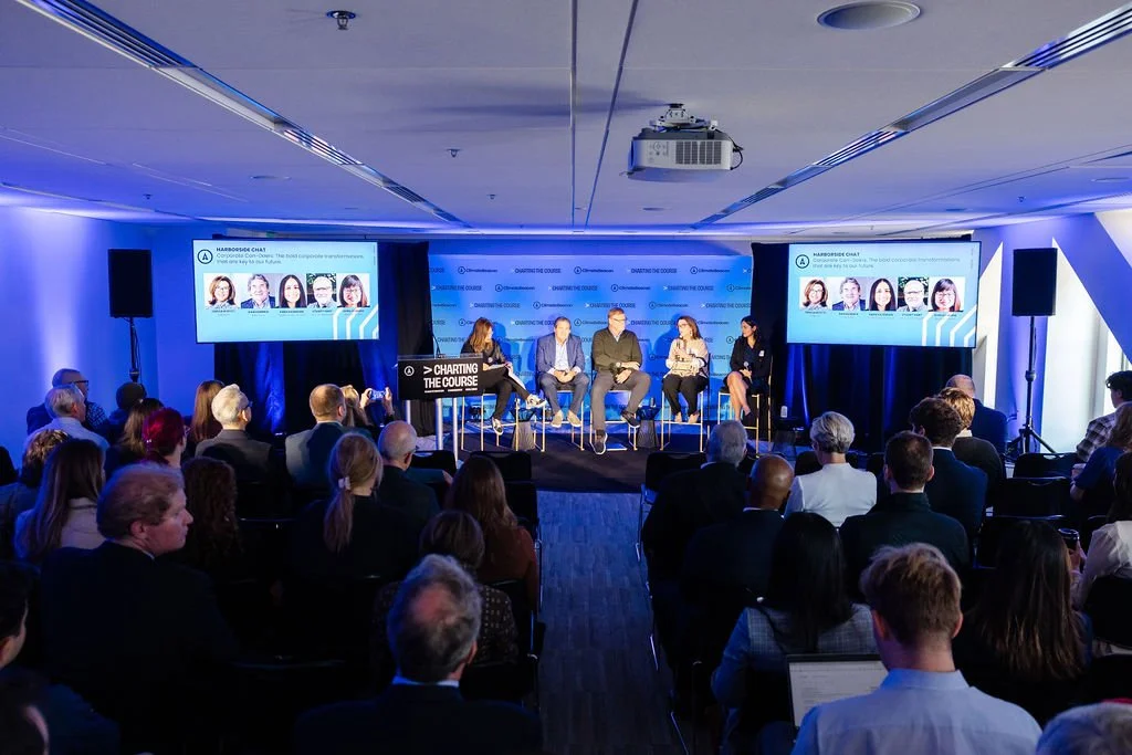 A conference panel discussion with five speakers on stage, two large screens displaying speaker images and text, and an audience seated in a darkened room with blue lighting.