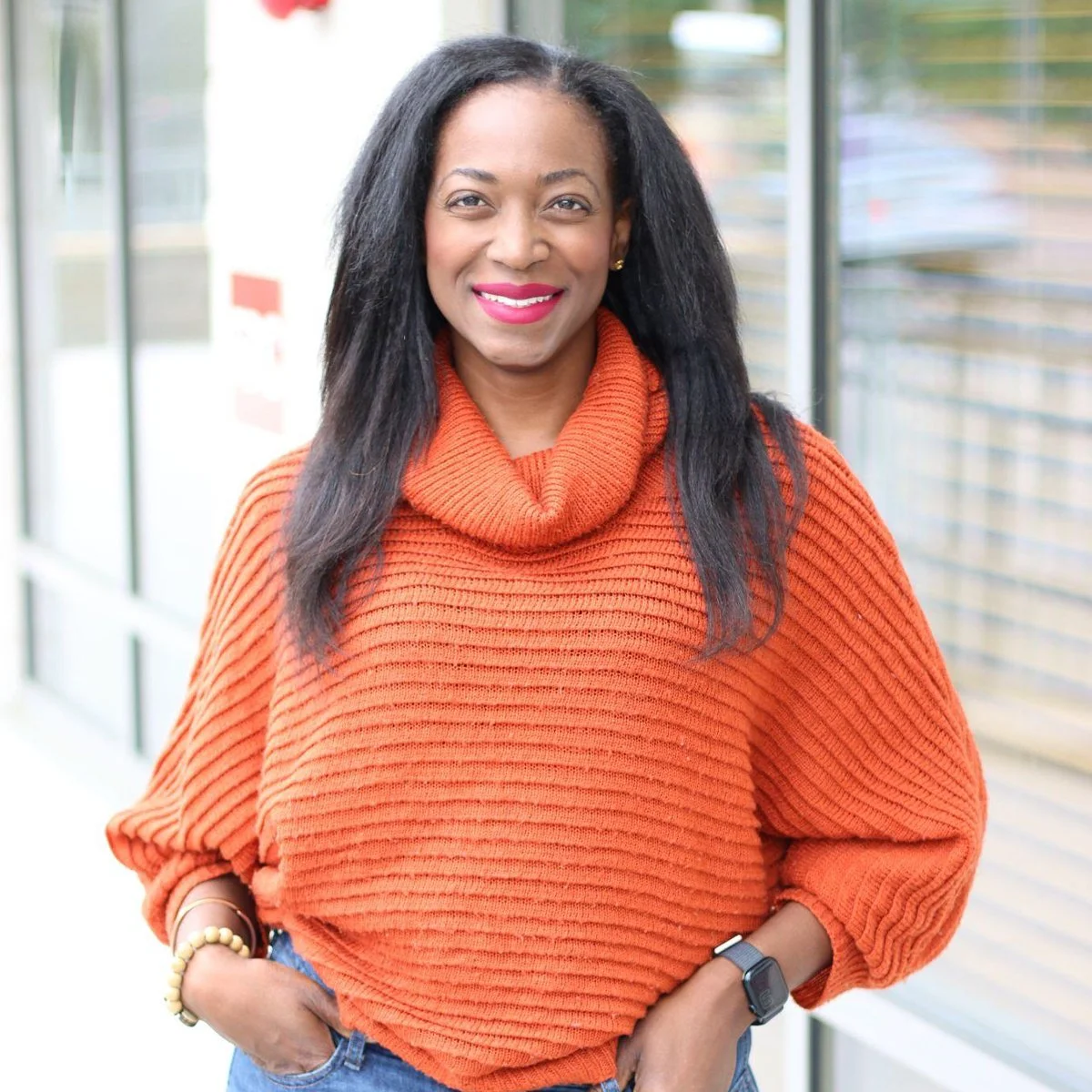 A woman with long black hair and wearing a bright orange ribbed turtleneck sweater, smiling outdoors in front of a glass building.