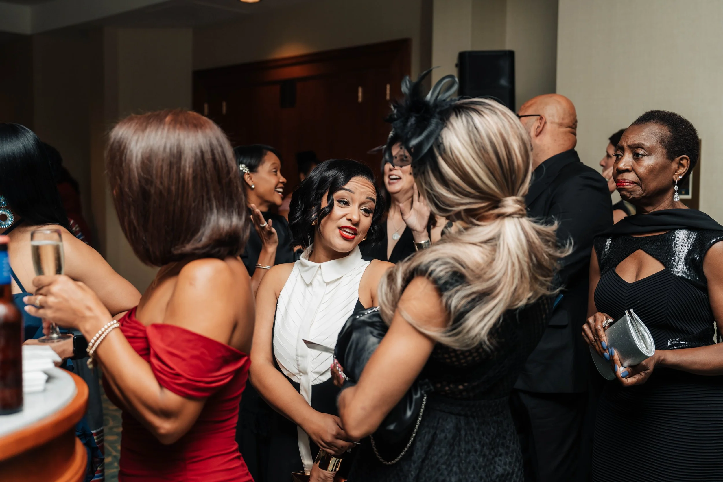Group of women dressed in elegant attire socializing at an indoor event, some holding glasses of wine or champagne.