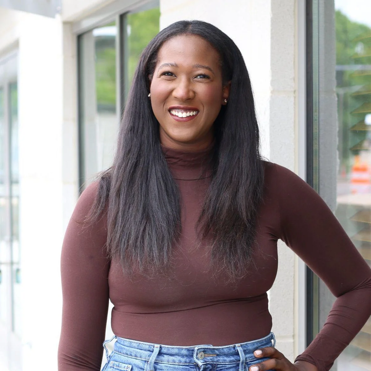 Portrait of a smiling woman with long black hair wearing a brown long-sleeve top and blue jeans, standing by a large window with green trees outside.