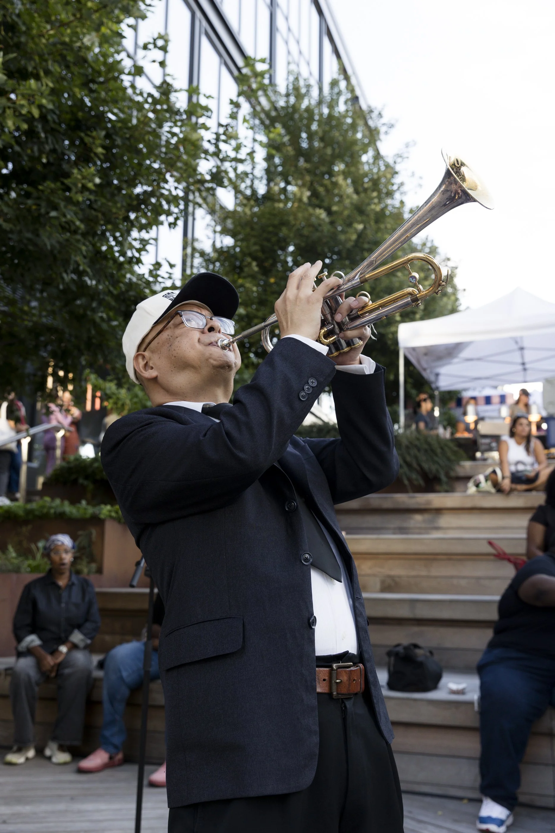 A man in a suit and glasses playing a trumpet outdoors, surrounded by people sitting on wooden steps and standing, with trees, a building, and a tent in the background.