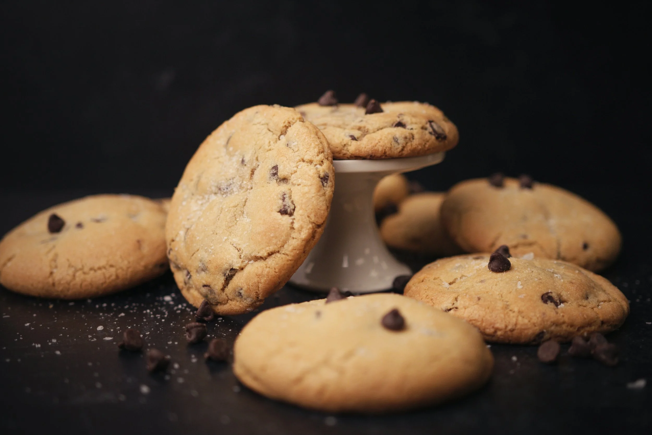 Chocolate chip cookies on a black surface with a white stand, some cookies leaning against the stand, sprinkled with salt and chocolate chips.
