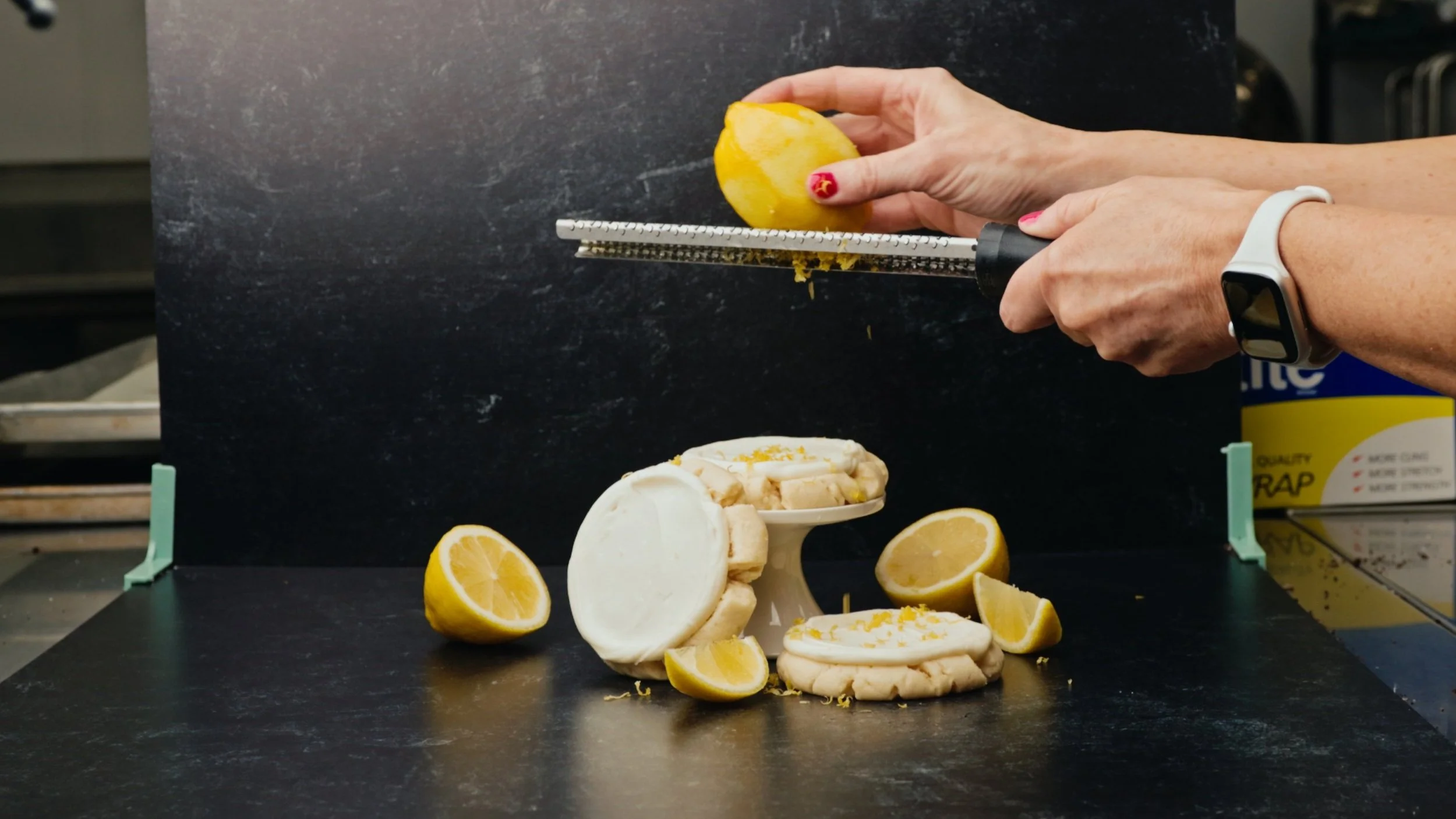 A person grating lemon zest over a plate of lemon-flavored meringue desserts, with lemon halves and wedges on a black surface.