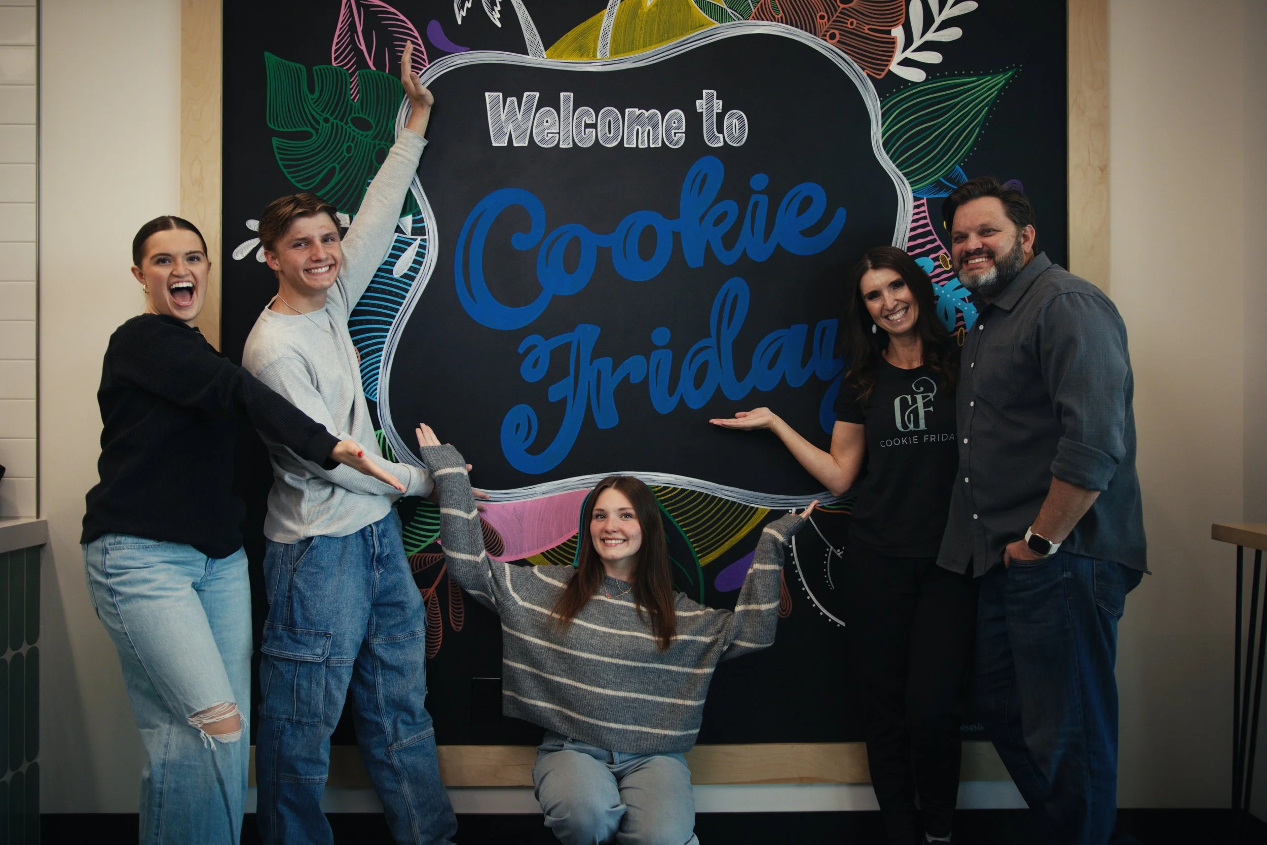 Group of five people smiling and posing in front of a decorated blackboard that reads 'Welcome to Cookie Friday' at a Cookie Frida event.