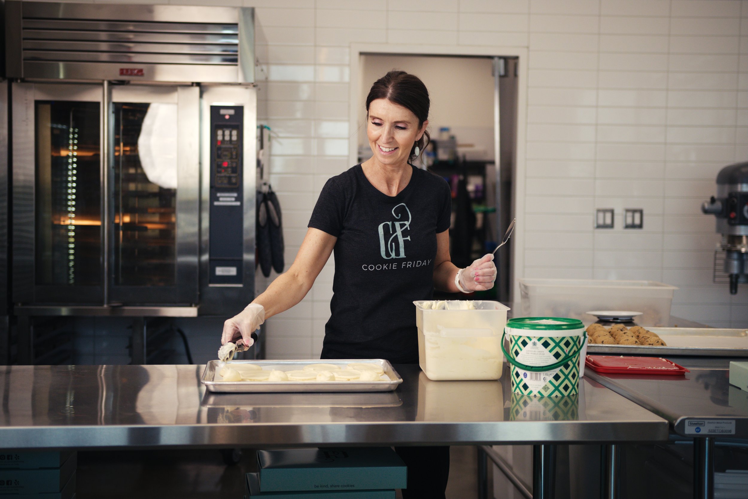 Woman in a bakery or kitchen preparing cookies on a stainless steel countertop, wearing a black t-shirt that says 'Cookie Friday', with baking supplies and cookies in the background.