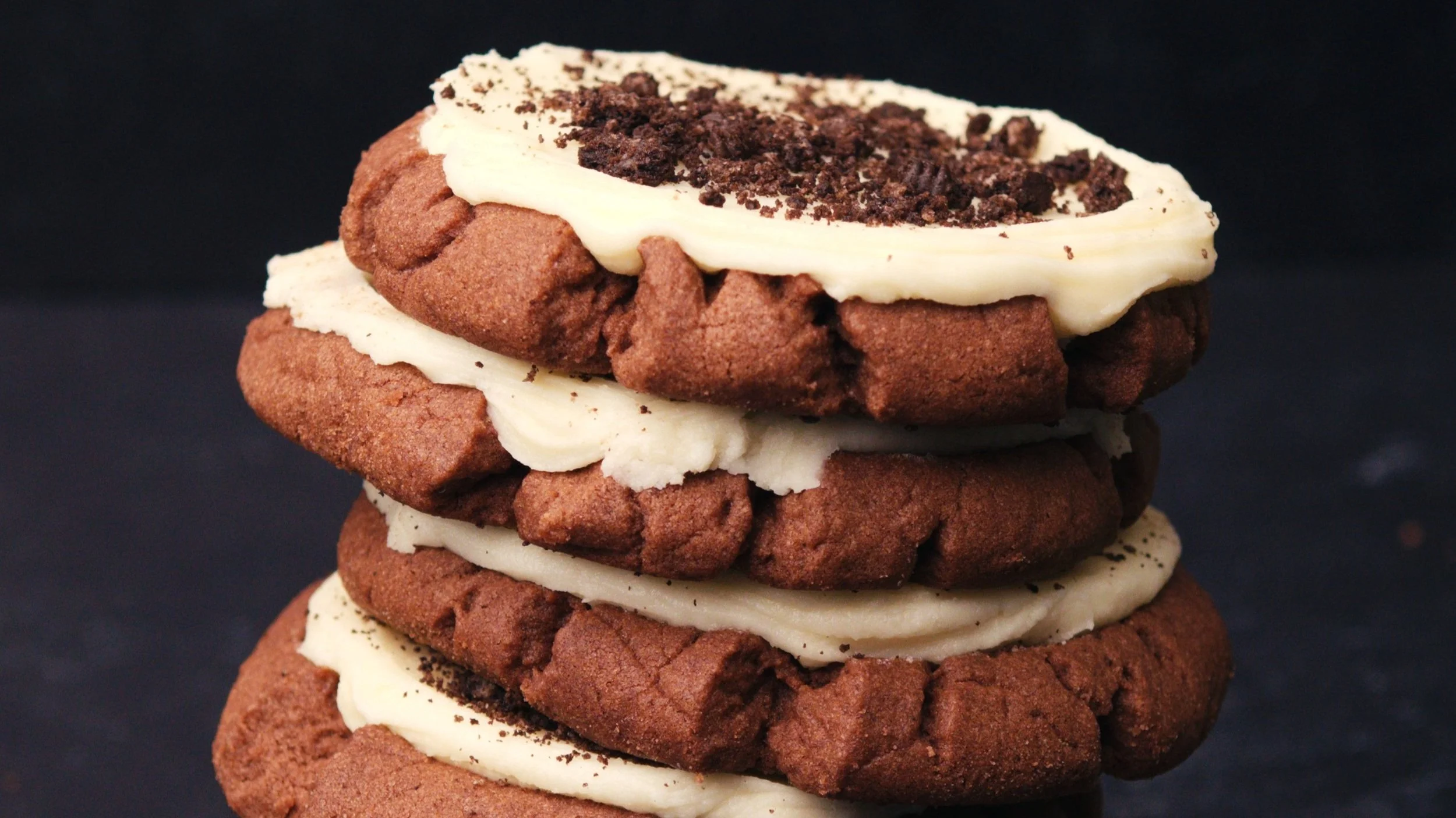 A stack of four chocolate cookies with vanilla frosting and chocolate crumbs on top against a black background.