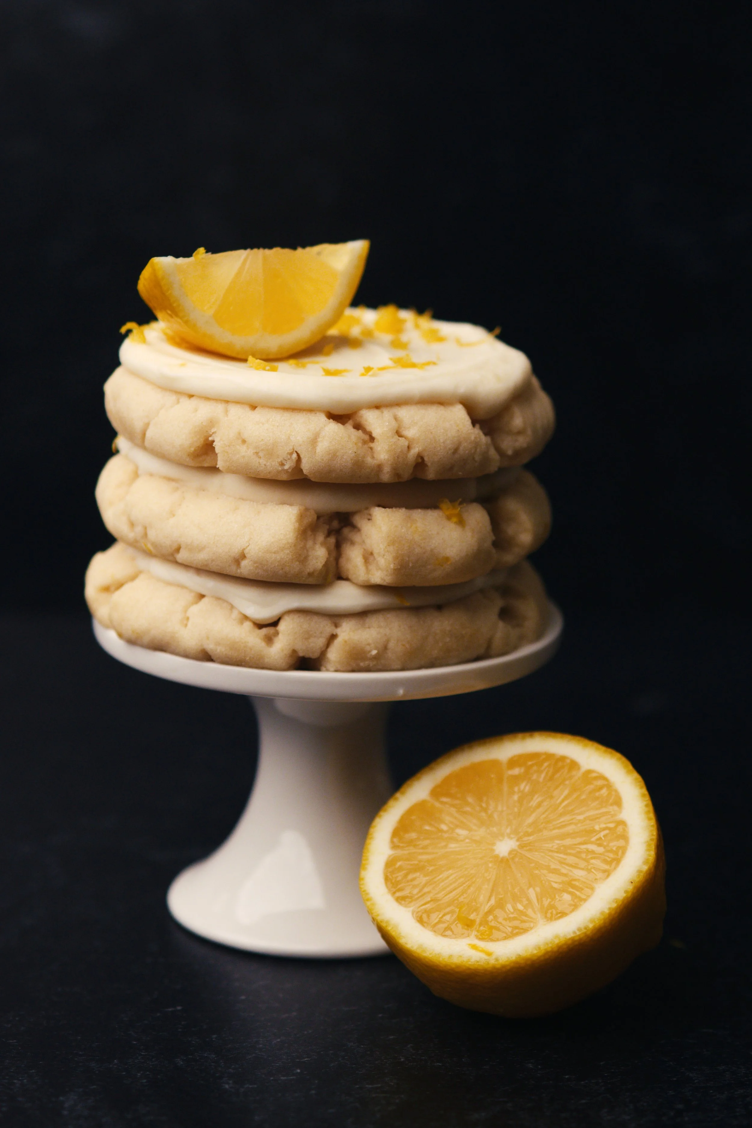 Lemon cookies with white frosting and lemon zest on a white cake stand, with a halved lemon in front, against a black background.