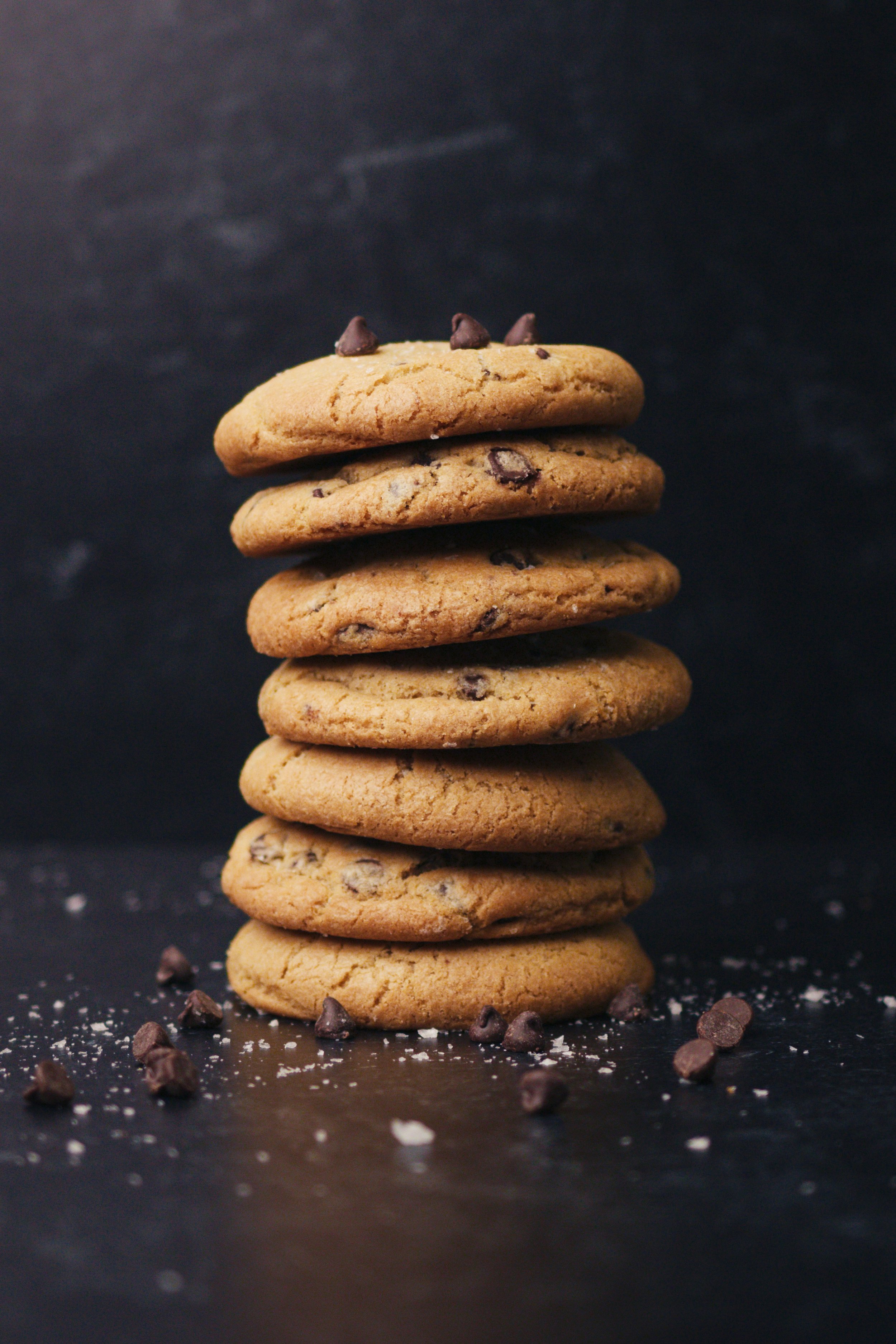 Stack of seven chocolate chip cookies, with some chocolate chips and crumbs scattered around, against a black background.