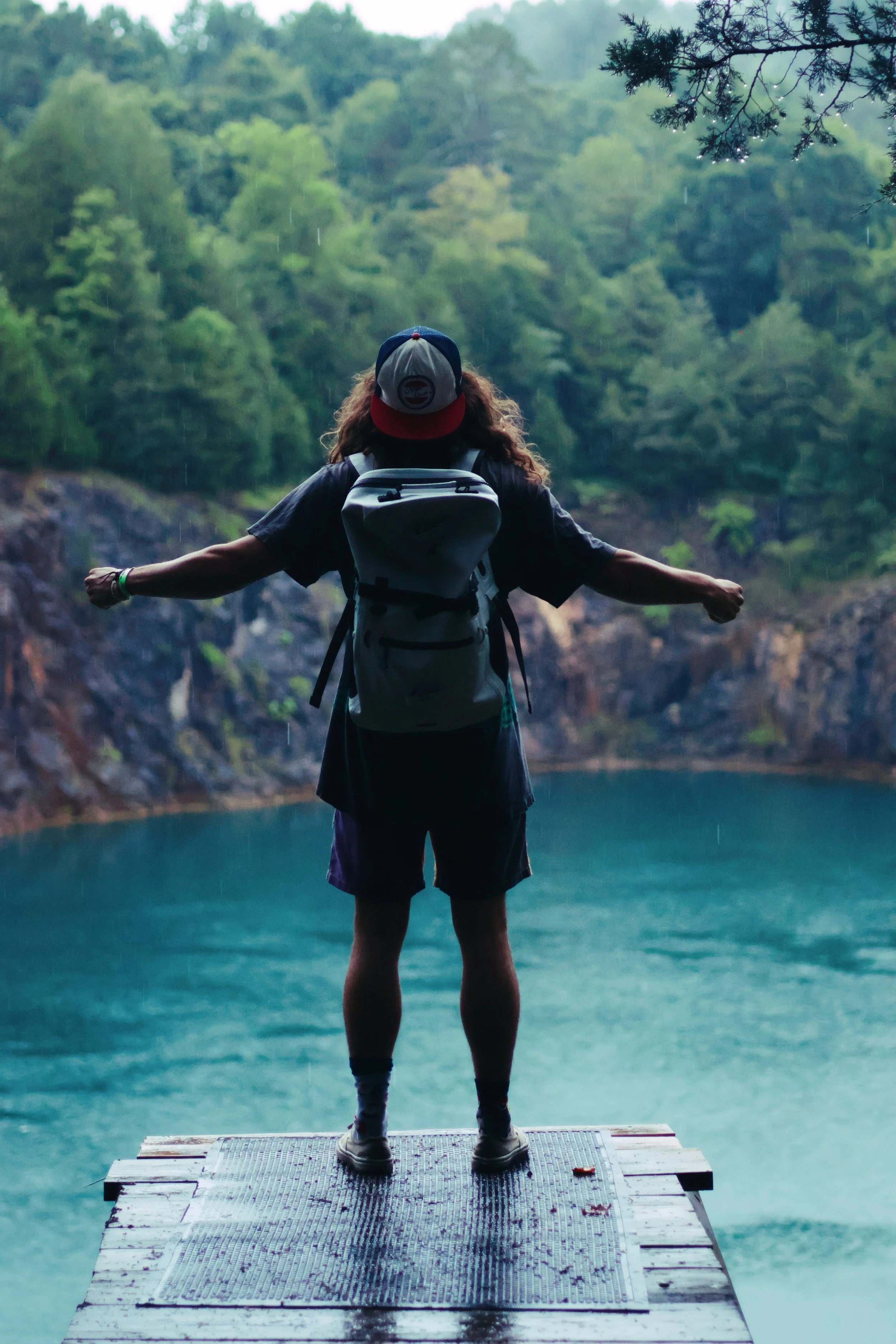 Person standing on a wooden dock with outstretched arms overlooking a lake, surrounded by dense green forest, with rain falling.