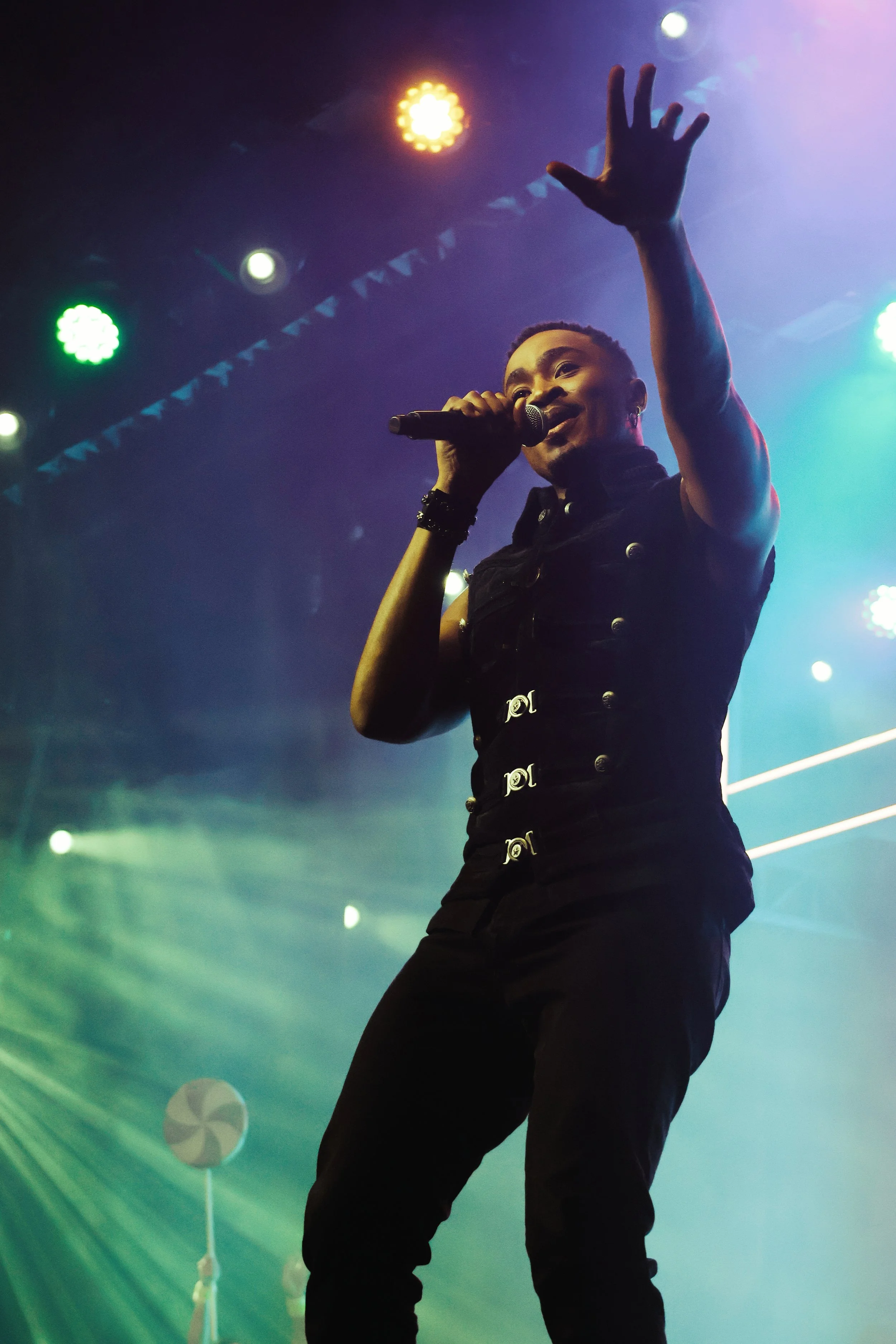 A performer singing on stage with a microphone, wearing a black sleeveless jacket with silver buttons, surrounded by colorful stage lights.