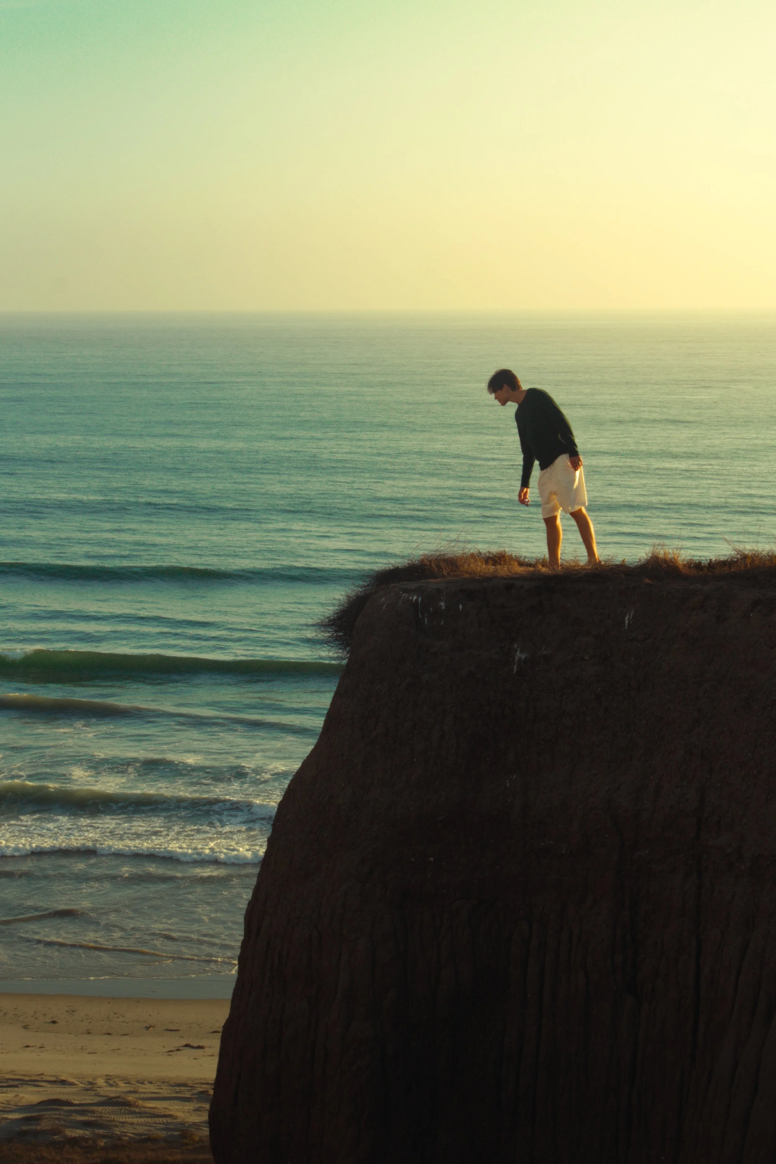 A young man standing on a cliff overlooking the ocean during sunset, wearing a black shirt and white shorts.
