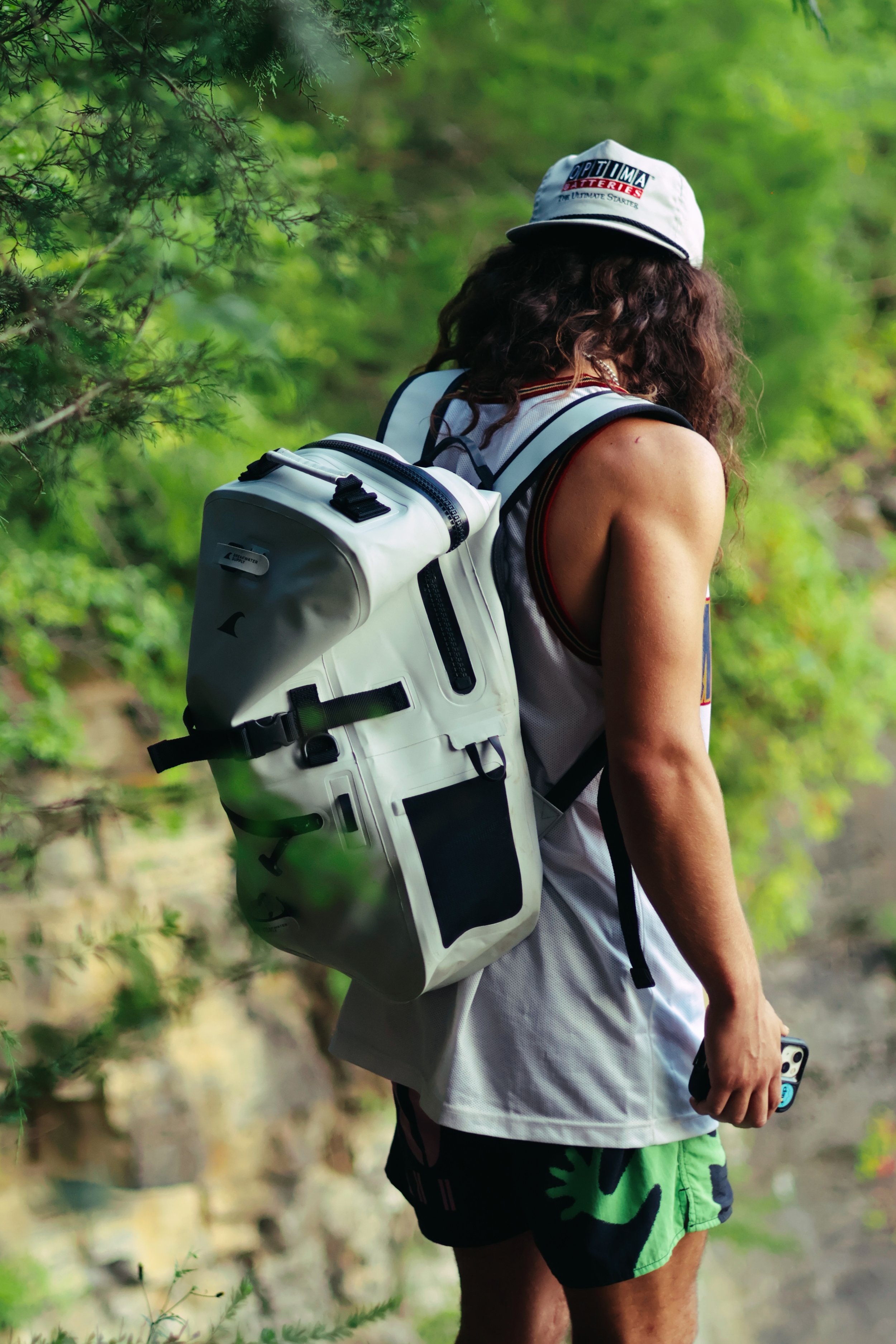 Person with curly hair wearing a white cap, sleeveless white top, and green and black shorts, carrying a large white backpack with black straps outdoors surrounded by green foliage.