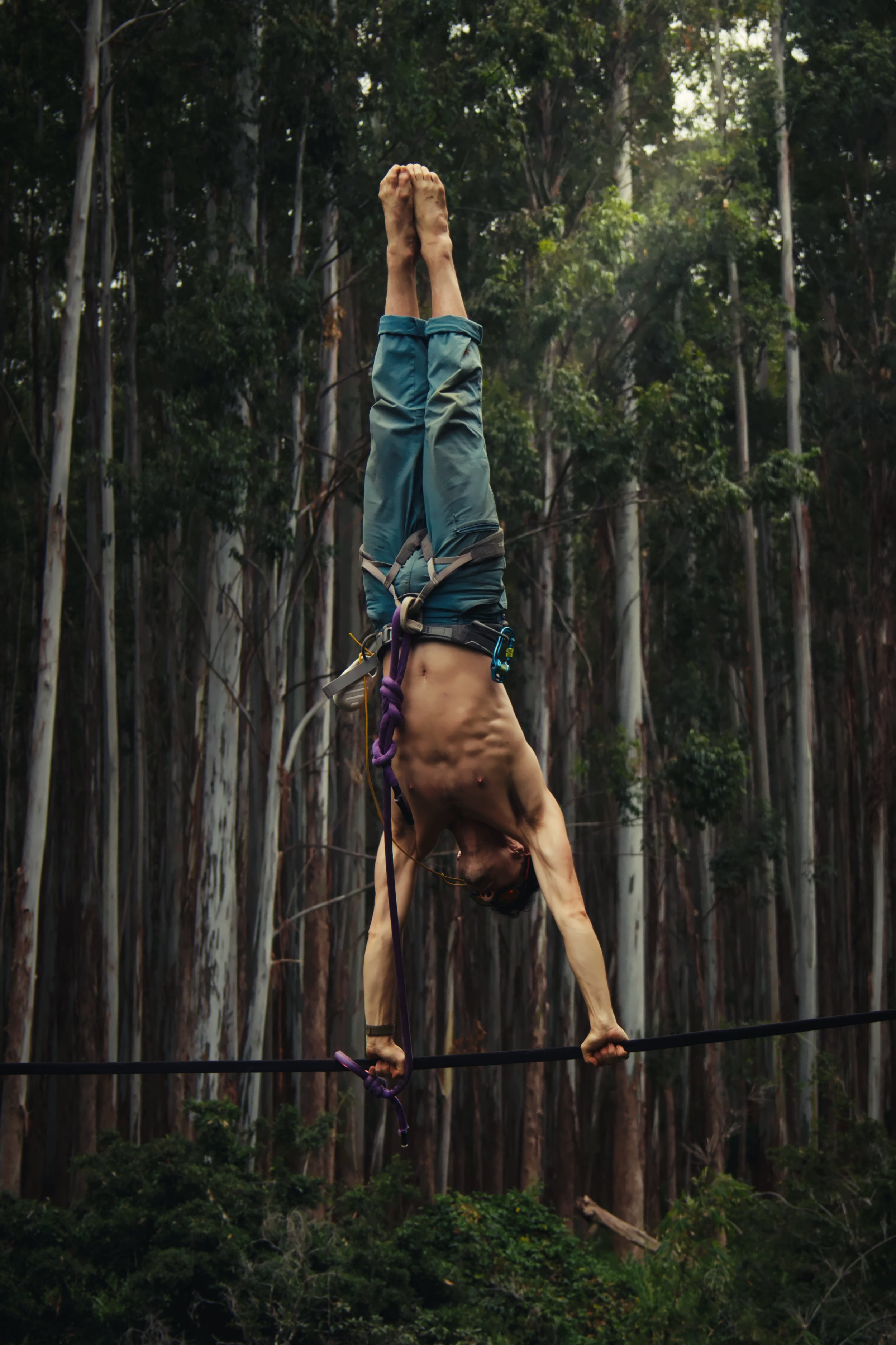 A man in climbing gear balancing upside down on a slackline between trees in a forest.