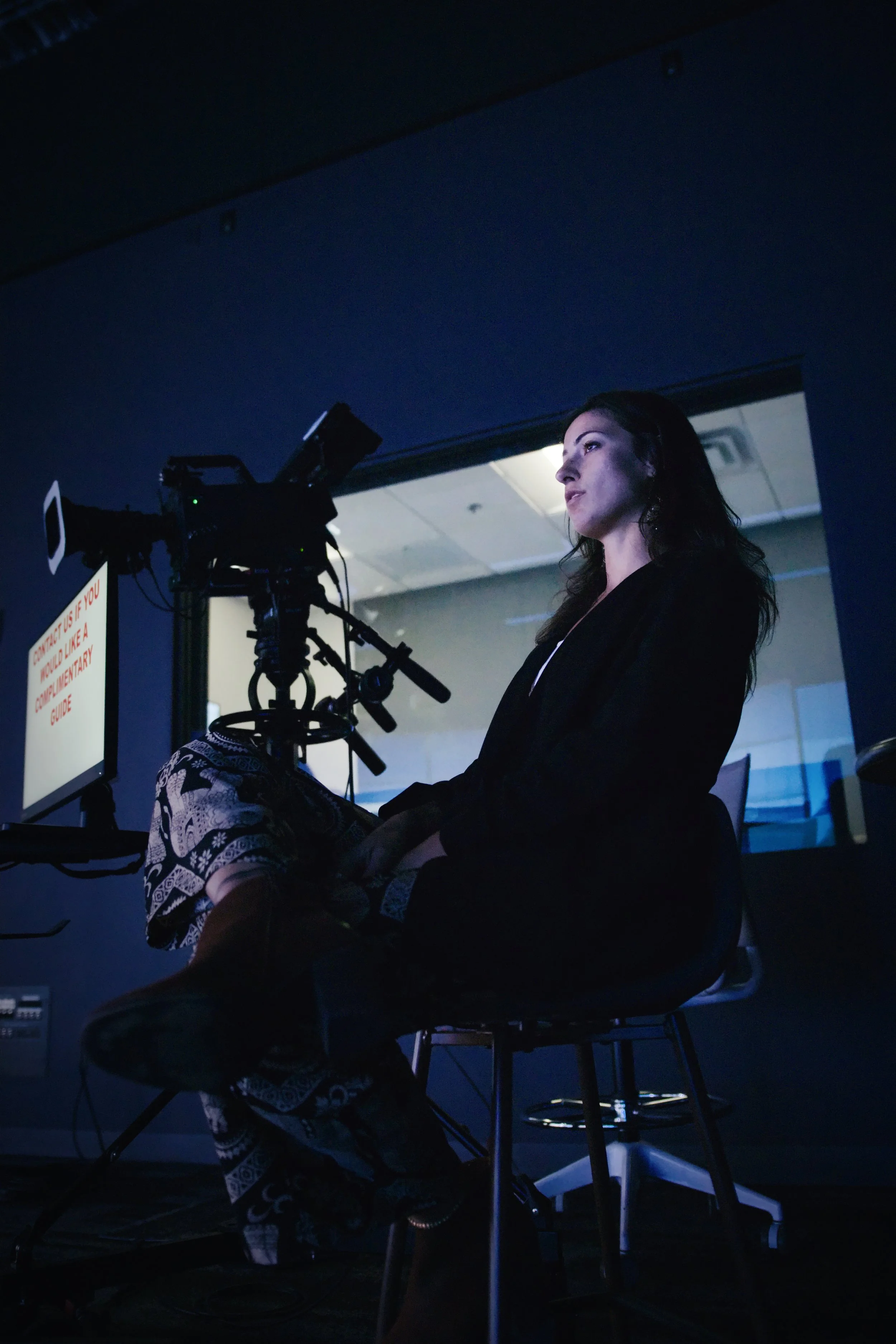 A woman sits in front of a camera in a dark room, with a monitor to her left displaying text and a lit backdrop behind her.